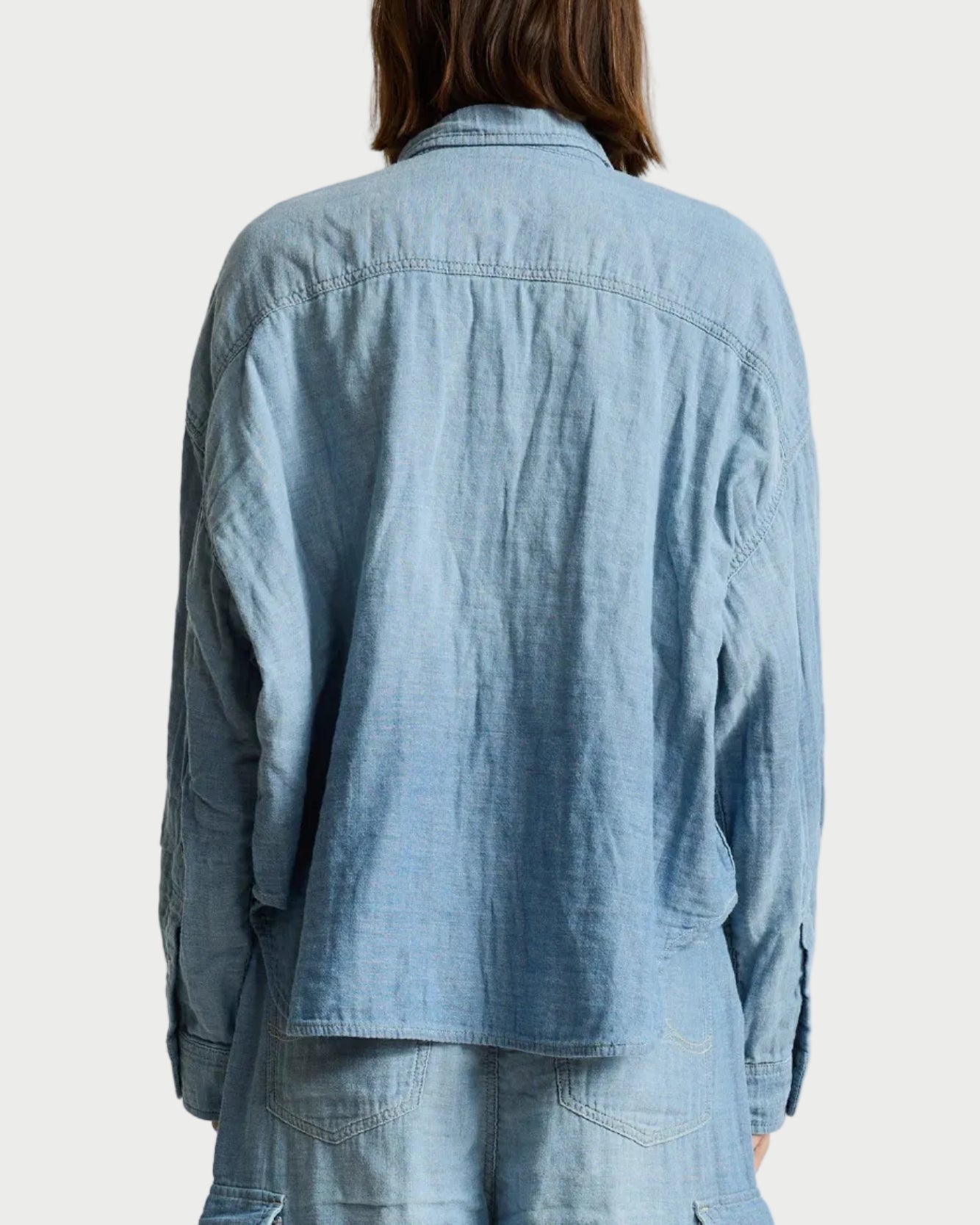 A person viewed from the back wearing a light blue denim shirt jacket and matching denim bottoms against a plain light gray studio background.