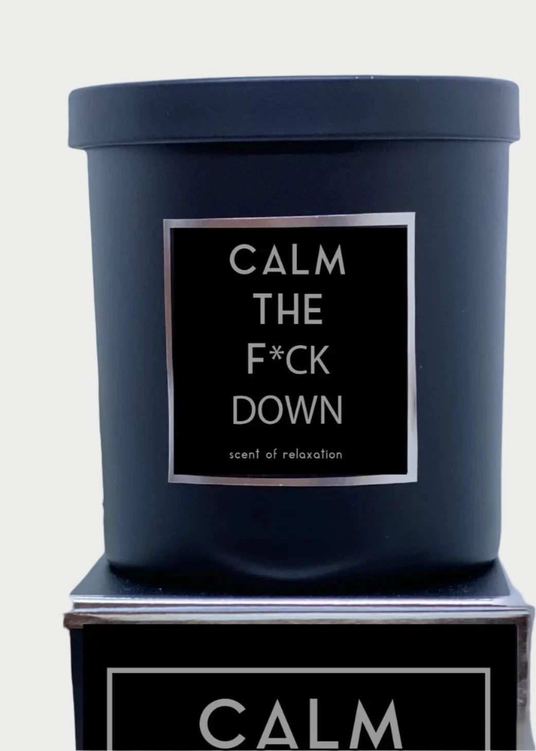A black 9oz candle with a label that reads "Calm the F*ck Down - scent of relaxation" is placed on a surface. Behind the Rude Dude Candles by Rude Dude, a metallic hand statue is visible, with one finger extended upward. Made in USA.