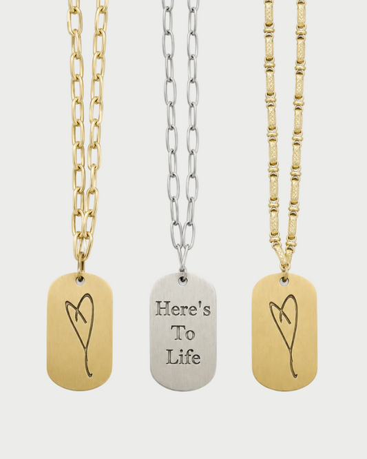Three dog tag necklaces displayed vertically on a light gray background. The left necklace has a gold chain and a gold tag engraved with a heart.