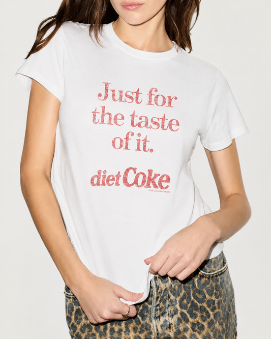 A woman models the RE/DONE Diet Coke Classic Tee, a relaxed fit cotton shirt by RE/DONE featuring vintage-inspired red lettering. She pairs it with leopard print pants and poses with one hand on her shirt.
