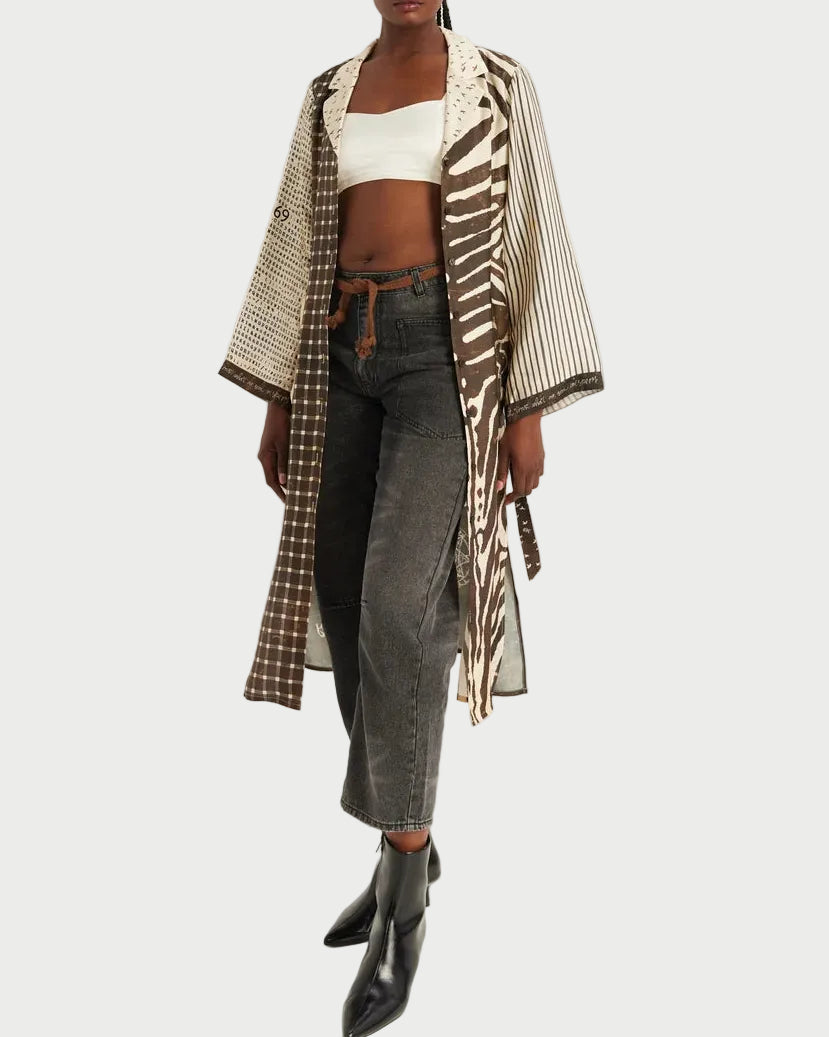 A model stands against a plain light background wearing a long open-front kimono-style jacket with mixed prints in beige, brown, and white, layered over a white crop top, dark gray jeans, and black ankle boots.