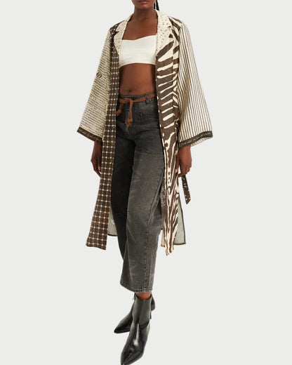 A model stands against a plain light background wearing a long open-front kimono-style jacket with mixed prints in beige, brown, and white, layered over a white crop top, dark gray jeans, and black ankle boots.
