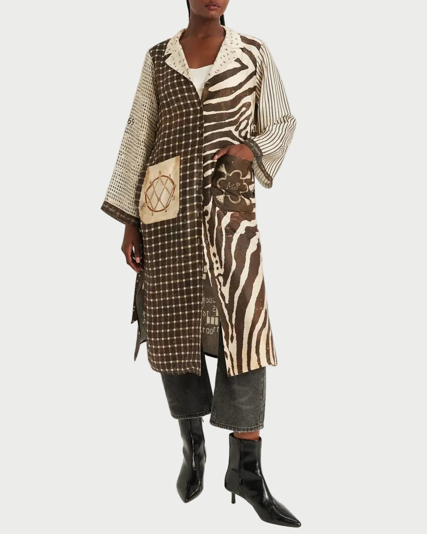 A model wearing a long, mixed-media patterned coat with brown and beige animal print, stripes, and checkered panels, styled with dark gray cropped pants and black pointed-toe ankle boots against a plain light gray studio background.