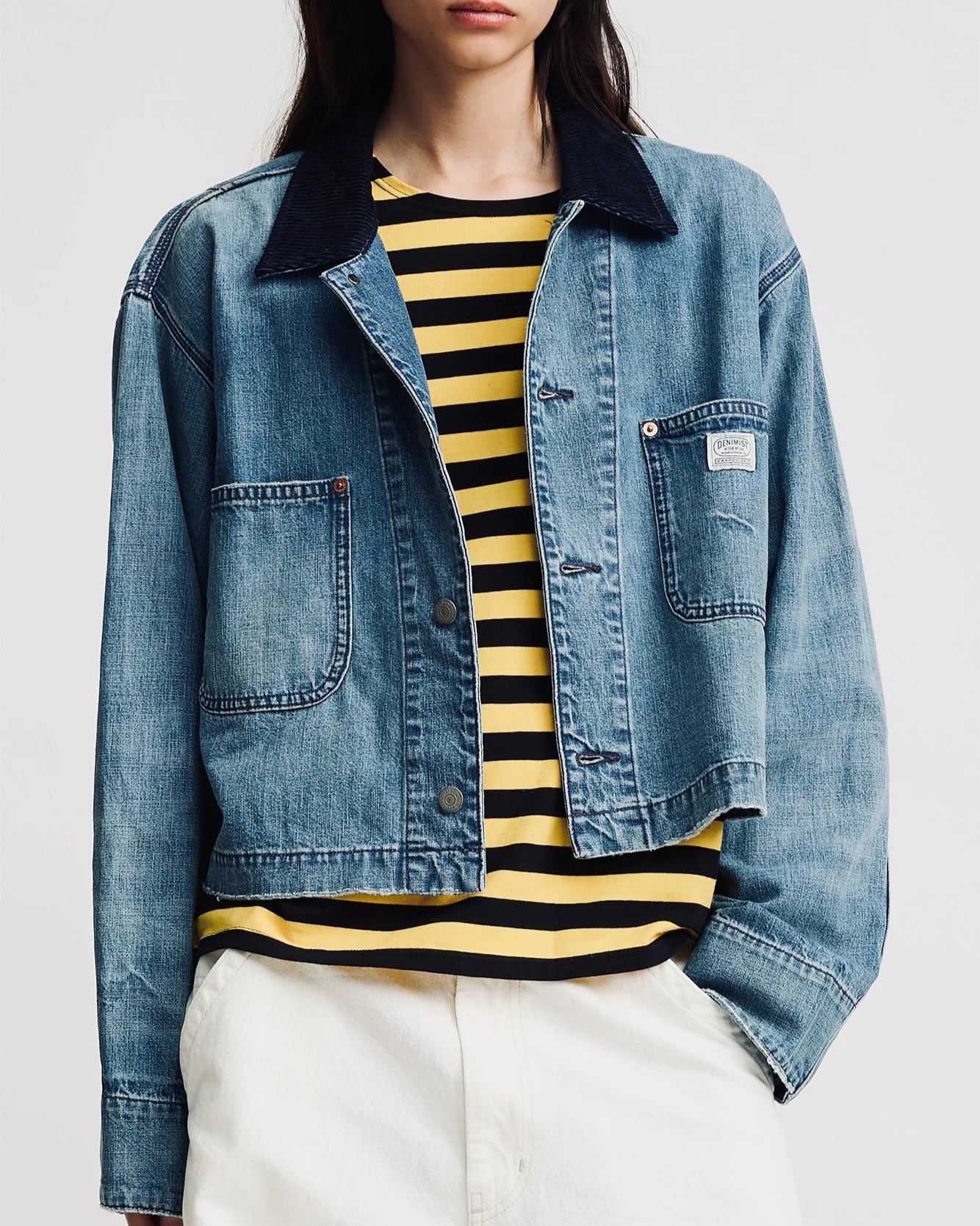 A person models the Denimist Chore Denim Jacket over a yellow and black striped shirt with white pants, posing with one hand in their pocket against a plain background.
