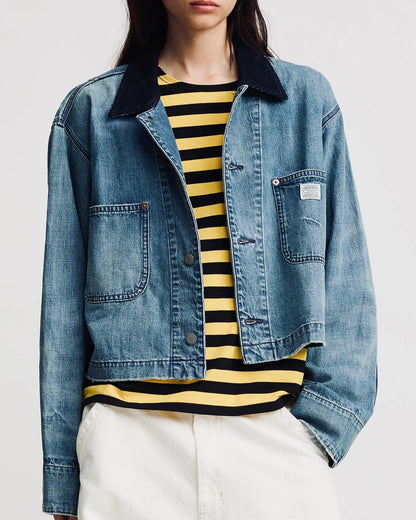 A person models the Denimist Chore Denim Jacket over a yellow and black striped shirt with white pants, posing with one hand in their pocket against a plain background.