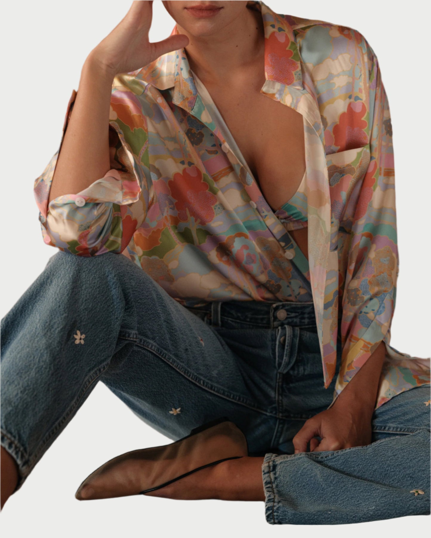 A person sits cross-legged wearing blue jeans, brown pointed flats, and WEAR CISSA's The Belle Shirt in a colorful Art Nouveau print, loosely buttoned over a matching top, with one hand resting on their face.