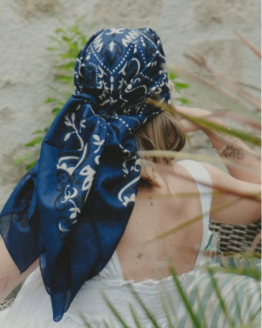 A woman with light brown hair sits outdoors, facing away. She wears the Maison Lecomte Flament Bandana Scarf Navy tied at the back and a white sleeveless dress, her right hand near her head as green plants blur in the foreground.
