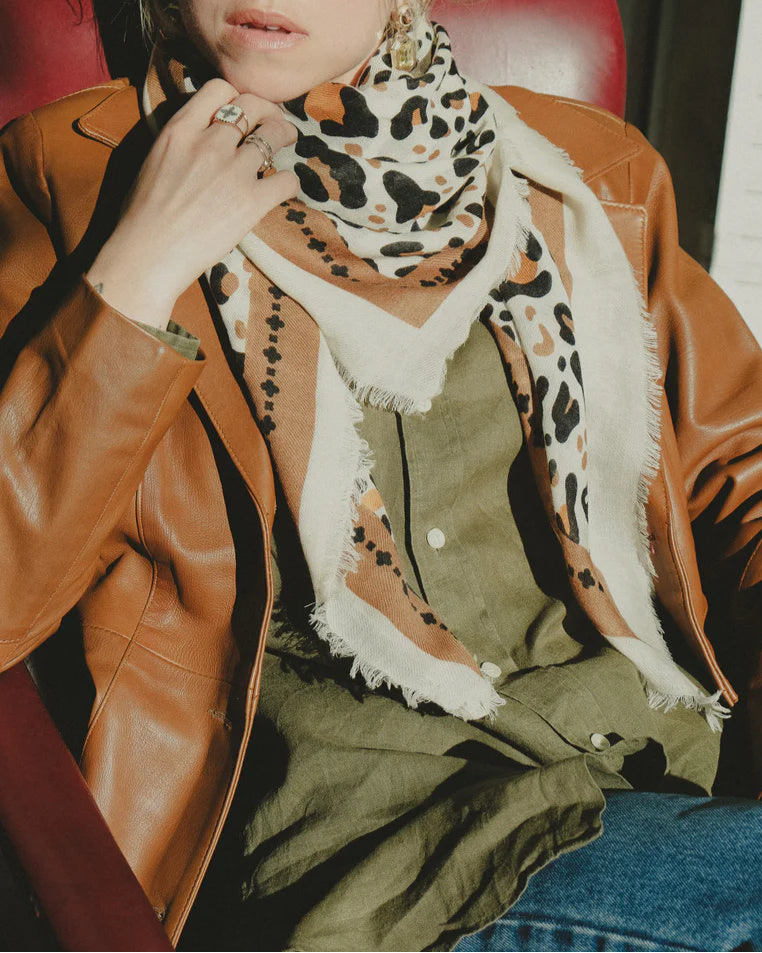 A person sits on a red chair wearing a brown leather jacket, olive green shirt, blue jeans, and the Maison Lecomte Flament Leo Scarf Beige. Their hand is near their face, displaying rings.
