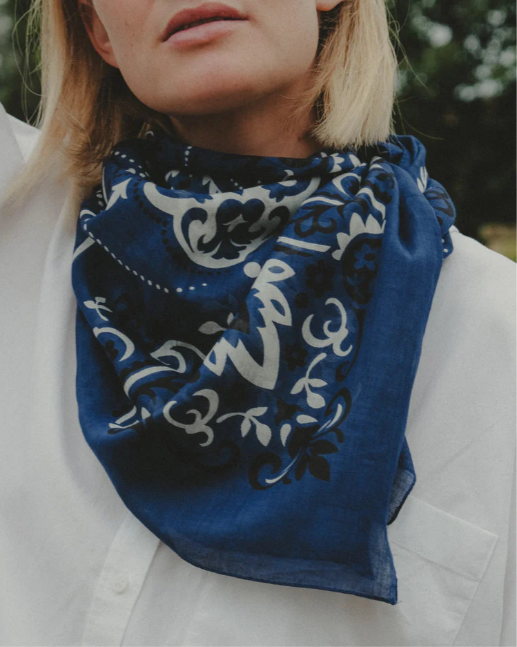 A person in a white shirt and a Maison Lecomte Flament Bandana Scarf Navy with a white and black paisley pattern, photographed from the shoulders up against a blurred greenery background.