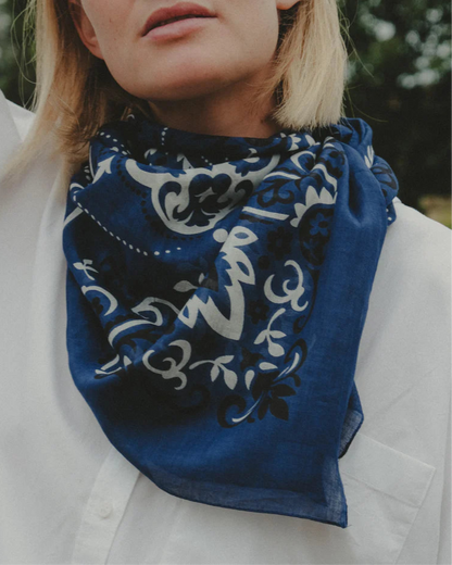 A person in a white shirt and a Maison Lecomte Flament Bandana Scarf Navy with a white and black paisley pattern, photographed from the shoulders up against a blurred greenery background.