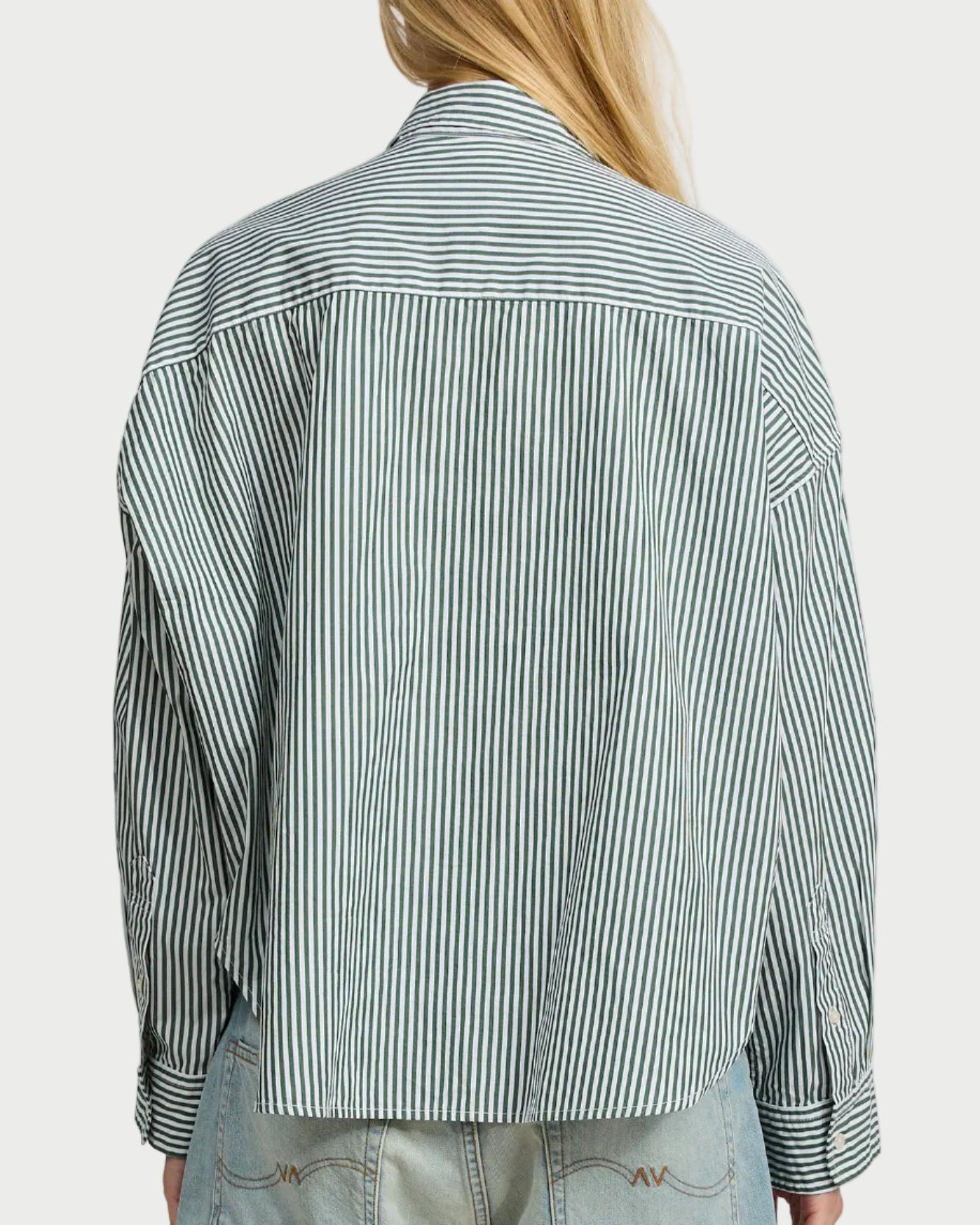 A person with long blonde hair, seen from the back against a white background, wears the Denimist Cropped Stripe Shirt by Denimist— a loose-fitting cotton button-down with black and white vertical stripes—paired with light blue jeans.