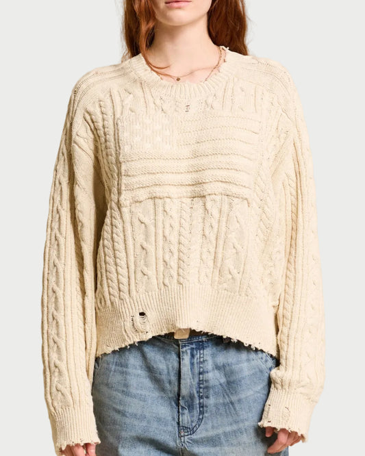 A person wears the Denimist Shadow Flag Sweater by Denimist—a loose, cream-colored premium cotton sweater with frayed edges and a small hole near the hem—paired with high-waisted blue jeans. The person’s face is partially out of frame.