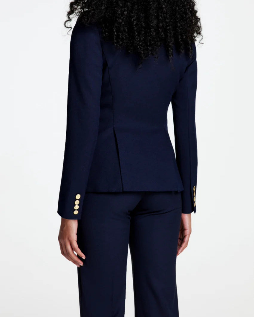 A person with curly hair, seen from behind, is wearing the Smythe Full Back Duchess Blazer in Navy with gold buttons and matching pants; the tailored cutaway silhouette of this blazer stands out against a plain white background.