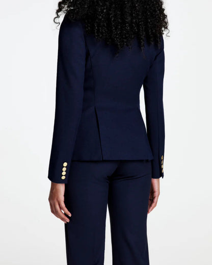 A person with curly hair, seen from behind, is wearing the Smythe Full Back Duchess Blazer in Navy with gold buttons and matching pants; the tailored cutaway silhouette of this blazer stands out against a plain white background.