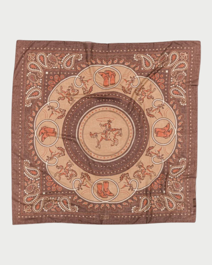 The Maison Lecomte Flament Rodeo Scarf Capuccino is a brown cotton voile scarf featuring a central cowboy on horseback, western motifs like boots, hats, saddles, horseshoes, and a paisley border.