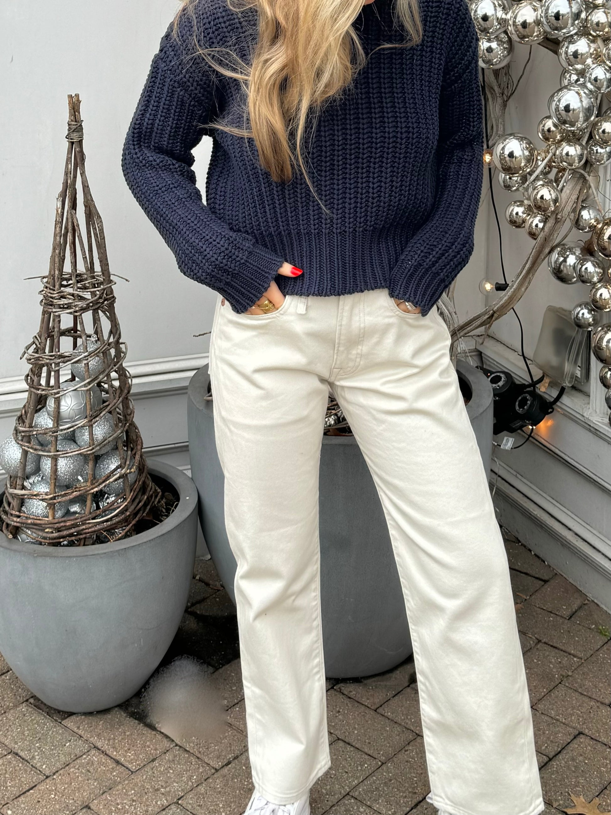 A person with long blonde hair stands outside next to silver holiday decorations and potted plants. They are wearing a chunky navy sweater and white high-waisted R13 Corduroy Boyfriend Jean with hands in the pockets. The background includes a silver bauble decoration and grey planters.