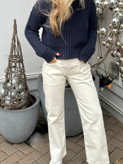 A person with long blonde hair stands outside next to silver holiday decorations and potted plants. They are wearing a chunky navy sweater and white high-waisted R13 Corduroy Boyfriend Jean with hands in the pockets. The background includes a silver bauble decoration and grey planters.