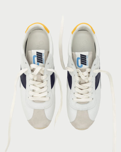 A top view of Oncept's Essex Sneaker shows sustainable design with black accents, white laces, yellow heel tabs, and blue and white tongue tags. The versatile footwear is set against a plain background.