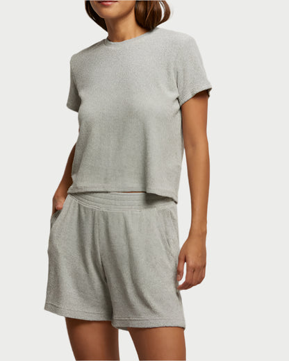 A person in a light gray, perfectwhitetee loungewear set called the Eve Loop Terry Tee, featuring a boxy-fit short-sleeve top and matching shorts. The casual outfit looks soft and offers ultimate comfort against a plain white background.
