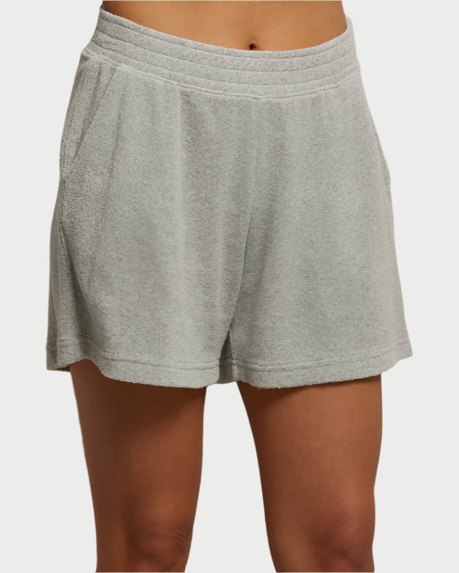 A person wearing Emily Loop Terry Shorts by perfectwhitetee, in light gray cotton with a soft loop terry texture. The shorts have an elastic waistband, side pockets, and reach knee-length. The background is plain and light-colored.