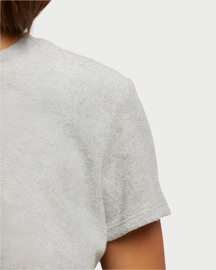 A close-up of a person wearing perfectwhitetee's Eve Loop Terry Tee emphasizes its light gray, textured loop terry fabric. The image highlights the shoulder and upper arm against a plain backdrop, showcasing the shirt's ultimate comfort in design.