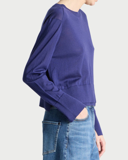 A person shown in side profile wearing a cropped long-sleeve knit top in a rich periwinkle blue-purple color, paired with blue jeans, against a clean light gray studio background.