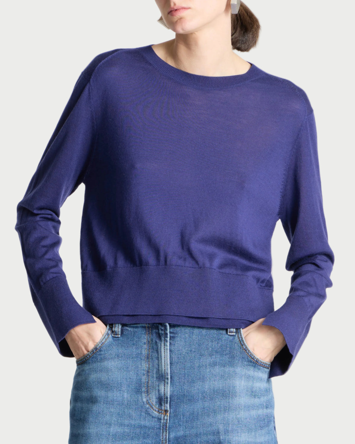A woman wearing a long-sleeve crewneck knit sweater in a deep periwinkle blue, paired with blue jeans, against a light gray studio background.