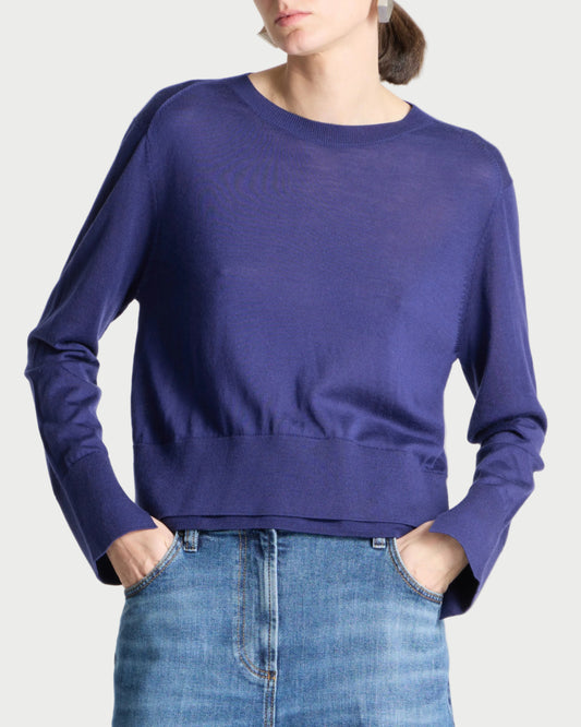 A woman wearing a long-sleeve crewneck knit sweater in a deep periwinkle blue, paired with blue jeans, against a light gray studio background.