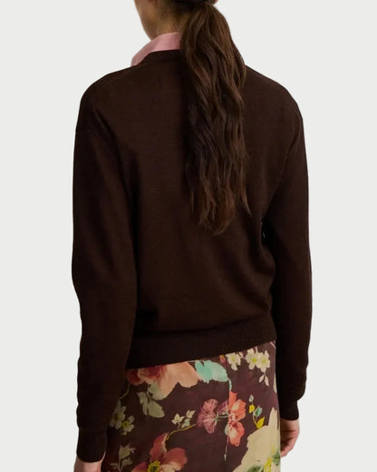 A woman viewed from behind wearing a dark brown long-sleeve knit sweater and a floral print skirt against a plain light gray background.