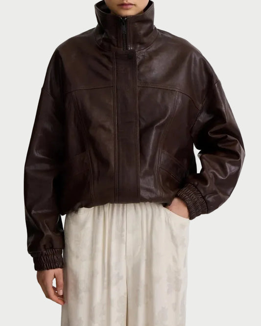 A person wears the Smythe Oversized Leather Bomber in dark brown leather with a high collar and elastic cuffs, paired with a light skirt. Their left hand is in the skirt pocket, face partly out of frame.