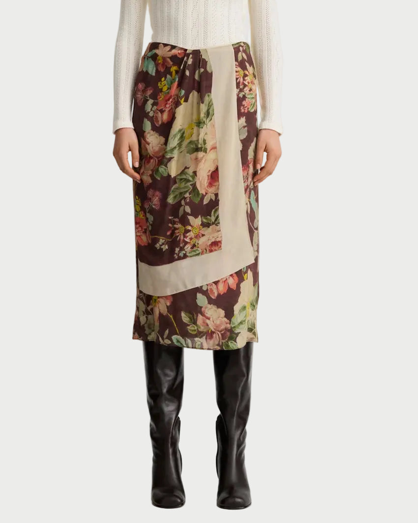 Wearing a white sweater, the model is styled in the Smythe Asymmetrical Overlay Skirt—a floral print midi skirt with a beige panel by Smythe—and black knee-high boots, shown from shoulders to feet.