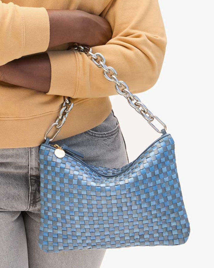 The person carries a handwoven blue leather Flat Clutch w/ Tabs by CLARE V., featuring a silver chain strap and a small round charm on the zipper, paired with a yellow sweater and light gray jeans.