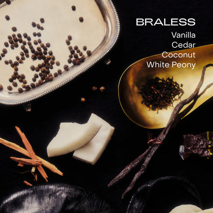 A flat lay of vanilla beans, cedarwood, coconut chunks, black peppercorns, and tea on black with the text: "BRALESS Vanilla Cedar Coconut White Peony" showcases the SIDIA Braless Candle, a non-toxic luxury home fragrance.