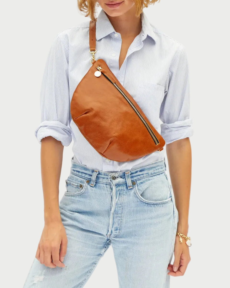 A person models the Clare V. Old Fashion Grande Fanny by CLARE V., a vegetable-tanned Italian leather fanny pack featuring a zipper and adjustable strap, worn stylishly across the chest with a light blue striped shirt and light-wash jeans.
