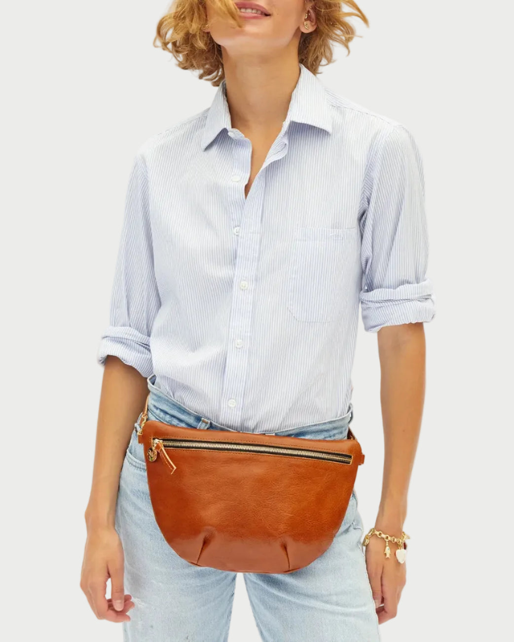 A woman wearing a light blue and white striped button-down shirt, light-wash jeans, and a brown leather belt bag with gold-tone hardware, posed against a clean white studio background.