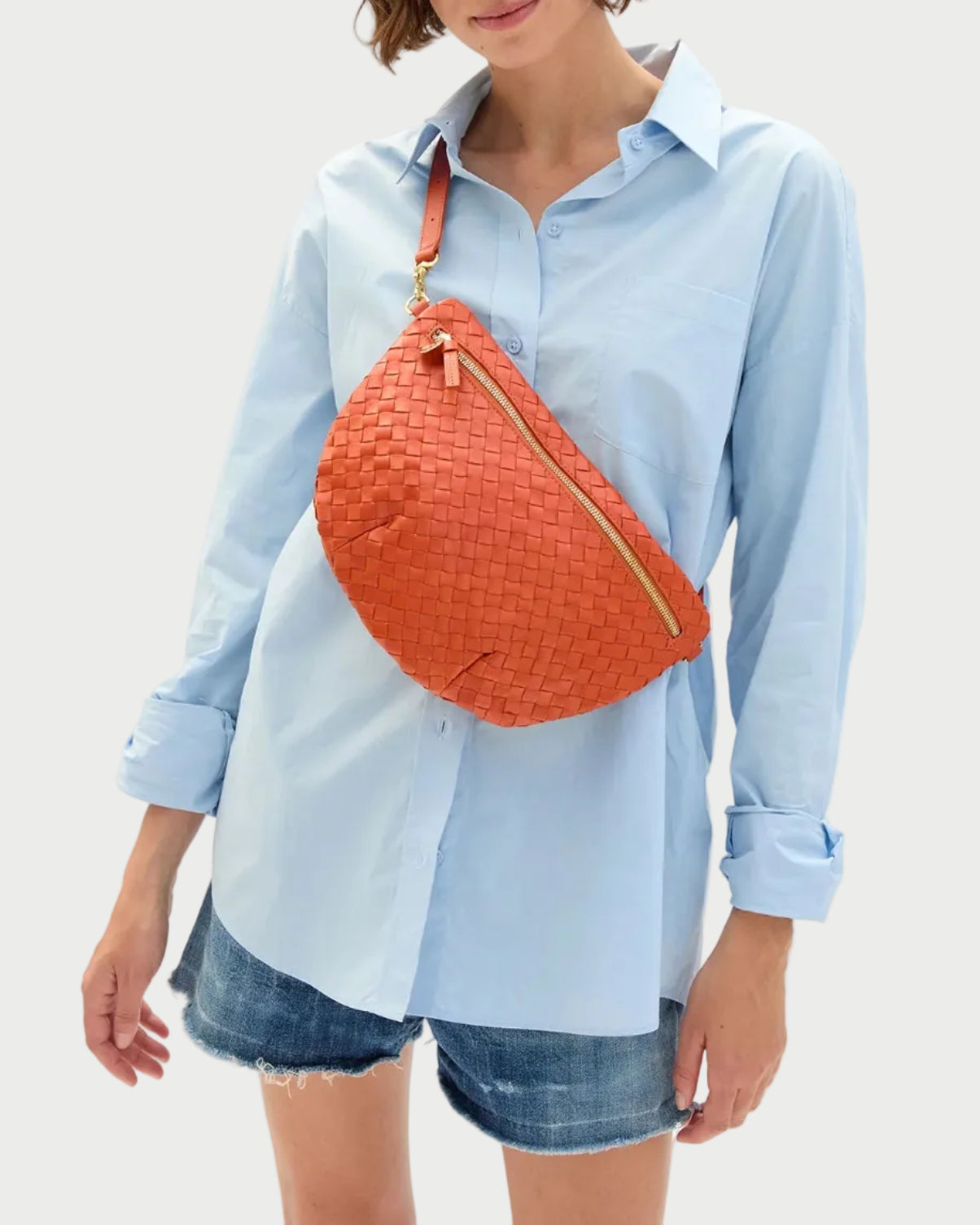 Woman in a light blue button-up shirt and denim shorts carries the CLARE V. Clare V. Woven Grande Fanny as a crossbody belt bag with a gold zipper across her chest.