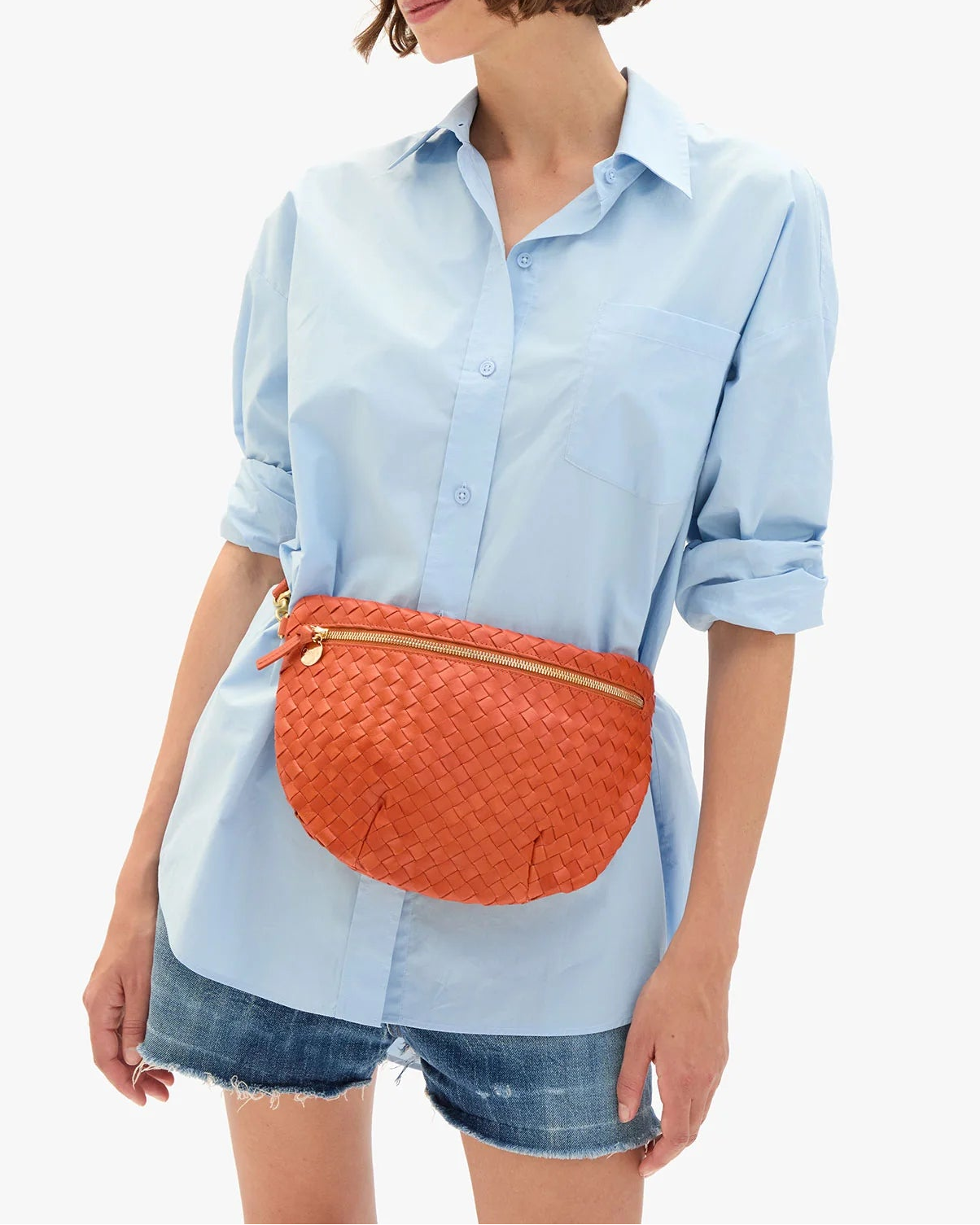 A woman in a light blue button-up shirt and denim shorts holds the CLARE V. Woven Grande Fanny, worn as a crossbody belt bag; her face is partially out of frame.