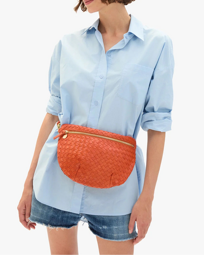 A woman in a light blue button-up shirt and denim shorts holds the CLARE V. Woven Grande Fanny, worn as a crossbody belt bag; her face is partially out of frame.