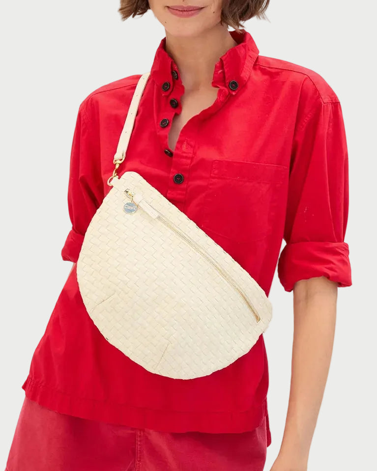 A woman in a bright red button-up shirt with rolled sleeves wears a large, cream woven Clare V. Grande Fanny by CLARE V. as a crossbody belt bag with a zipper.