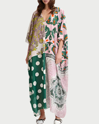 A woman wears the Hansas Printed Caftan by Essentiel Antwerp, featuring a loose, oversized fit with a deep V-neck and placed prints of florals, green and white polka dots, plus pink, green, and yellow paisley. She pairs it with white shoes.