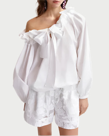A woman wears the Essentiel Antwerp Haquiri Top, an off-the-shoulder white blouse with ruffles and a large bow, paired with white floral-embroidered shorts featuring an elasticated waist.