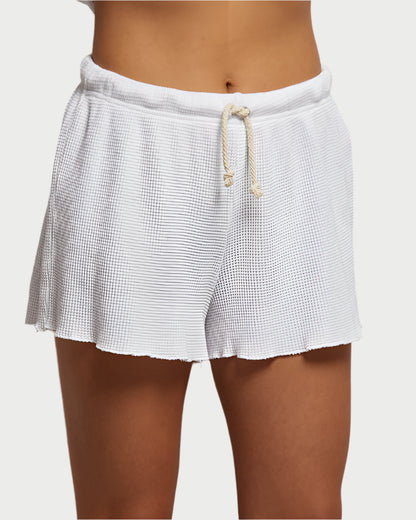 A person wearing Romy Waffle Shorts from perfectwhitetee stands against a plain background, highlighting the cozy waffle knit texture and stylish white drawstring. The subtle pattern enhances their laid-back look, perfectly paired with a casual Perfect White Tee.