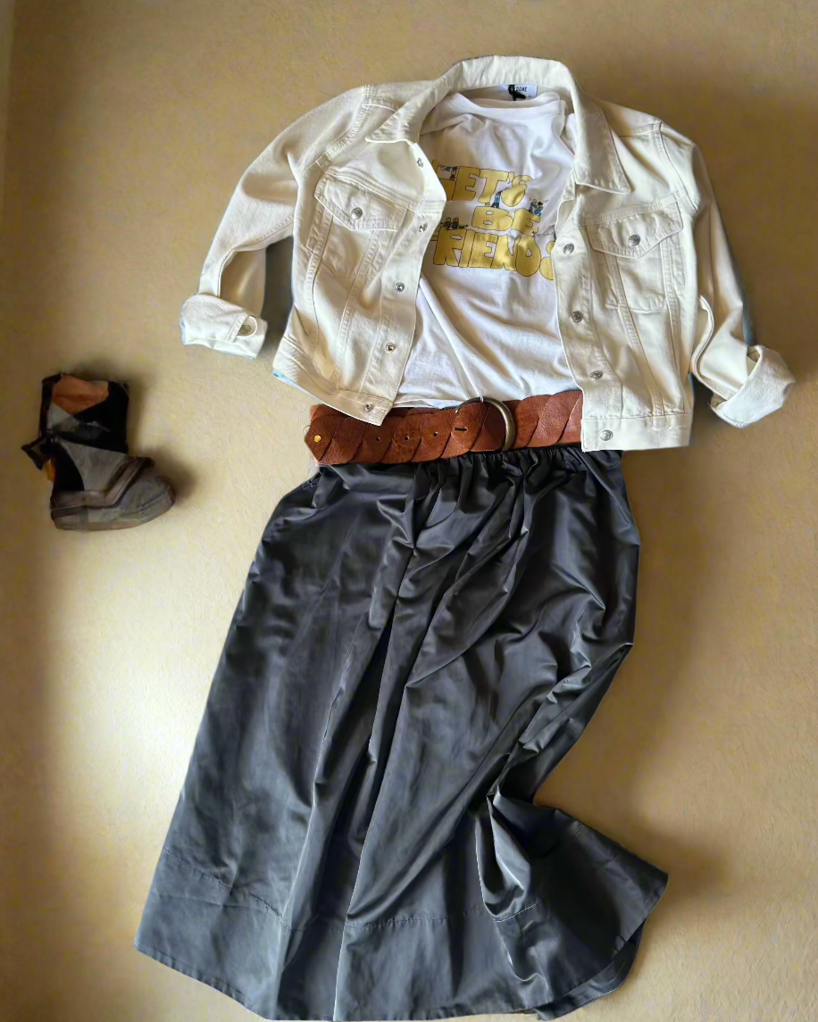 A casual outfit is displayed on a beige background, featuring a white denim jacket, white graphic tee, brown belt, the Miss Skirt by Herskind in gray, and brown ankle boots placed to the side.