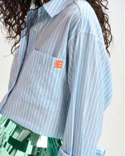 A person with long, wavy hair wears Essentiel Antwerp's blue and white Double Cuff Shirt with an orange logo on the chest pocket, paired with a shiny green paneled skirt.