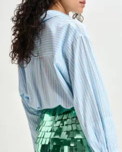 A person with long curly hair wears Essentiel Antwerp's Double Cuff Shirt, an oversized light blue striped cotton top, paired with a green metallic skirt featuring square sequin details, shown from side and back.
