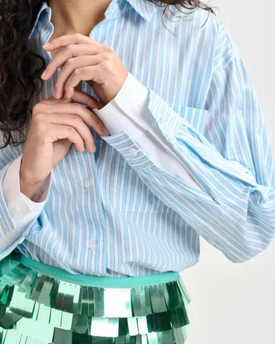 A person wears the Essentiel Antwerp Double Cuff Shirt, an oversized blue and white striped cotton button-up with white cuffs. Their face is not visible, and their hands are held near their chest.