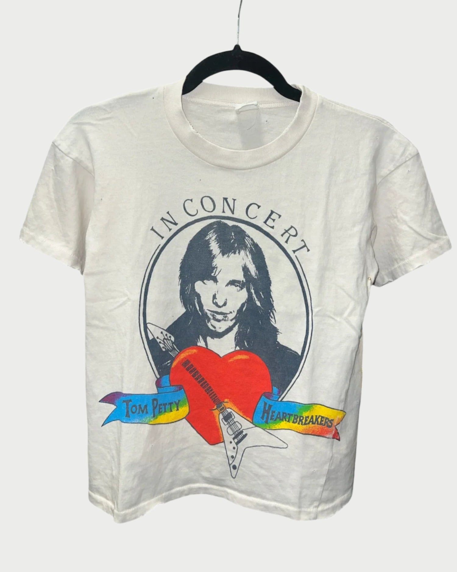 The Madeworn Tom Petty Tee, with its vintage fit, is a stylish 100% cotton white shirt featuring a graphic of a smiling face in a circle labeled "IN CONCERT." Beneath it is "Tom Petty Heartbreakers," surrounded by colorful banners and an electric guitar.