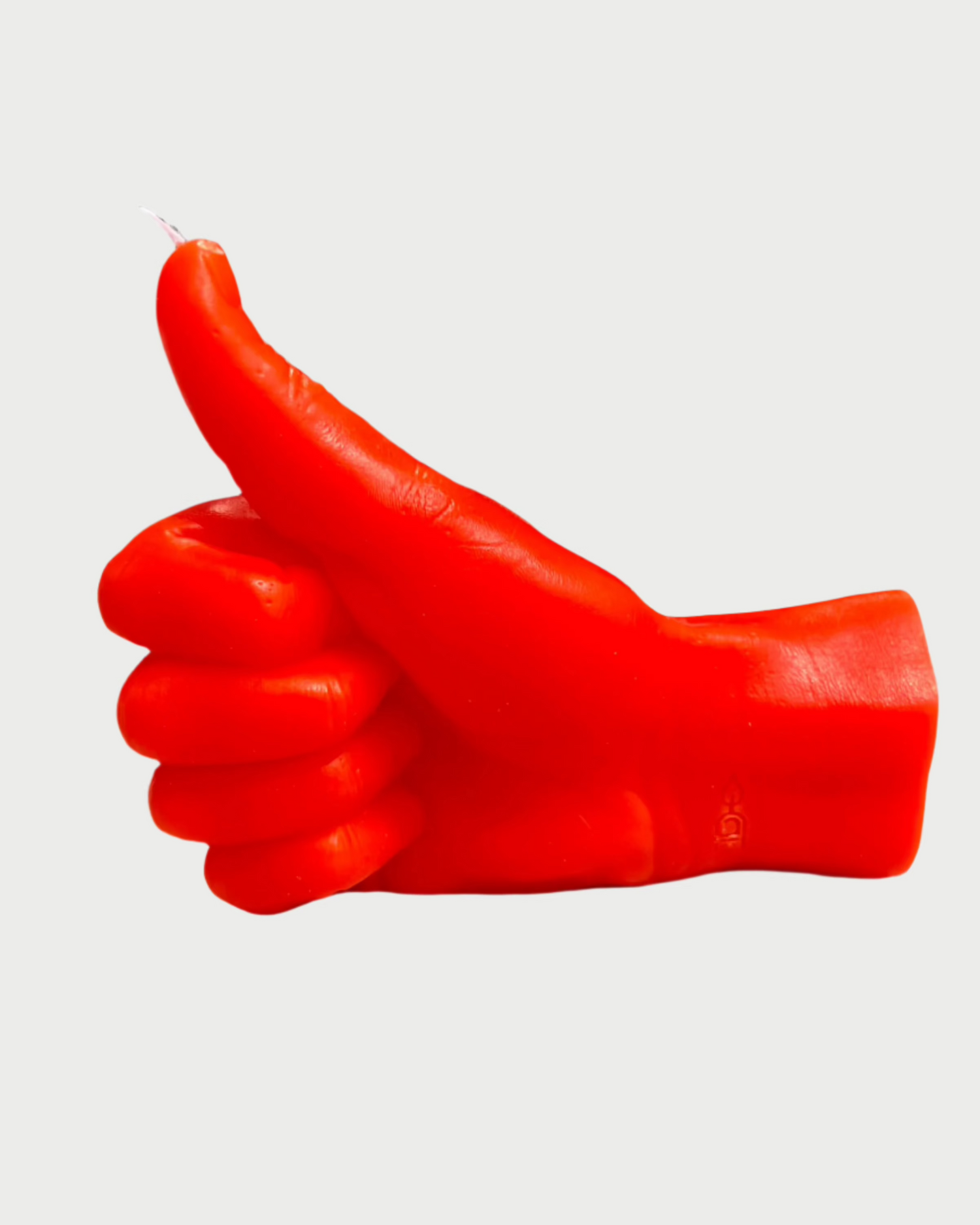 The 54° Celsius Thumbs Up Candle, a vibrant red hand-shaped candle on a light gray surface, vividly displays its color and character. Molded to depict a thumbs-up gesture, it stands out against the slightly blurred outdoor backdrop.