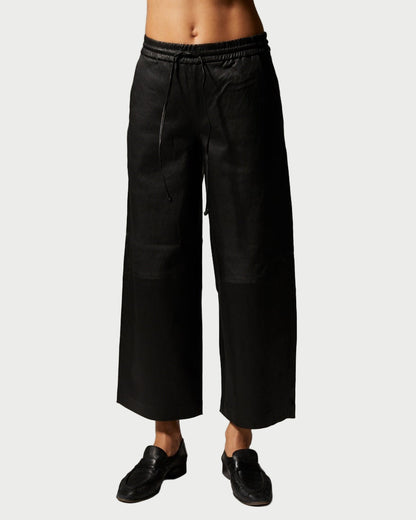 A person wears SPRWMN Cropped Leather Baggy Pants—black, wide-leg with an elastic waistband—paired with black shoes, standing against a plain light background.