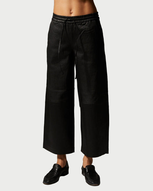 A person wears SPRWMN Cropped Leather Baggy Pants—black, wide-leg with an elastic waistband—paired with black shoes, standing against a plain light background.